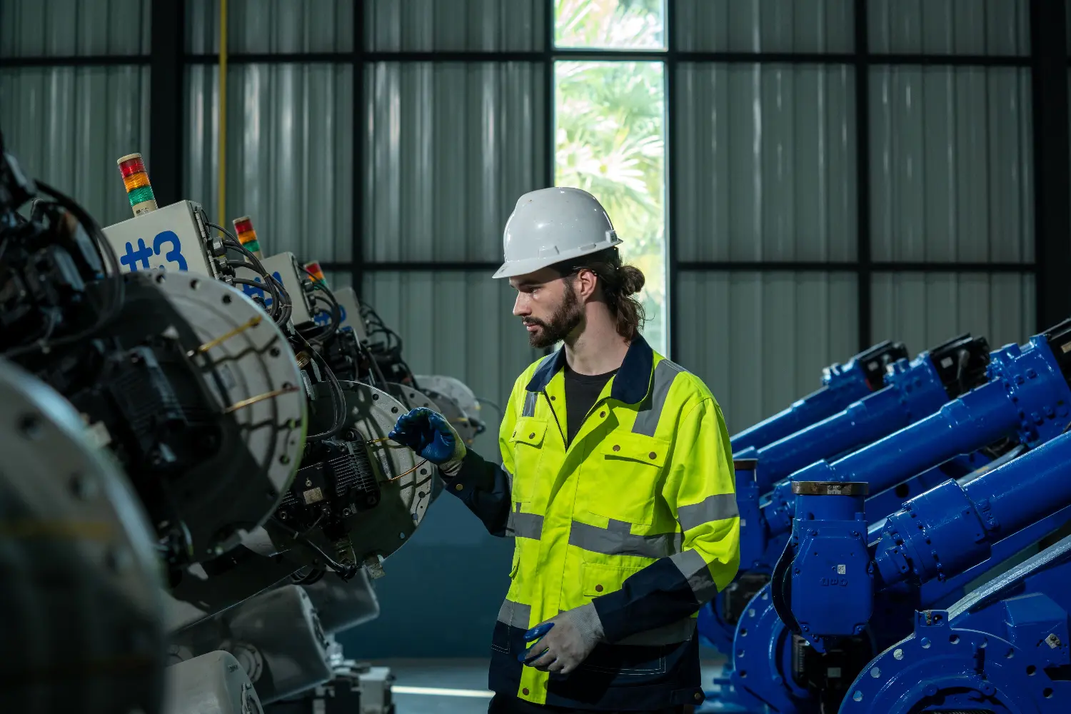 Técnico de segurança com capacete e EPI inspeciona robôs industriais azuis em fábrica
