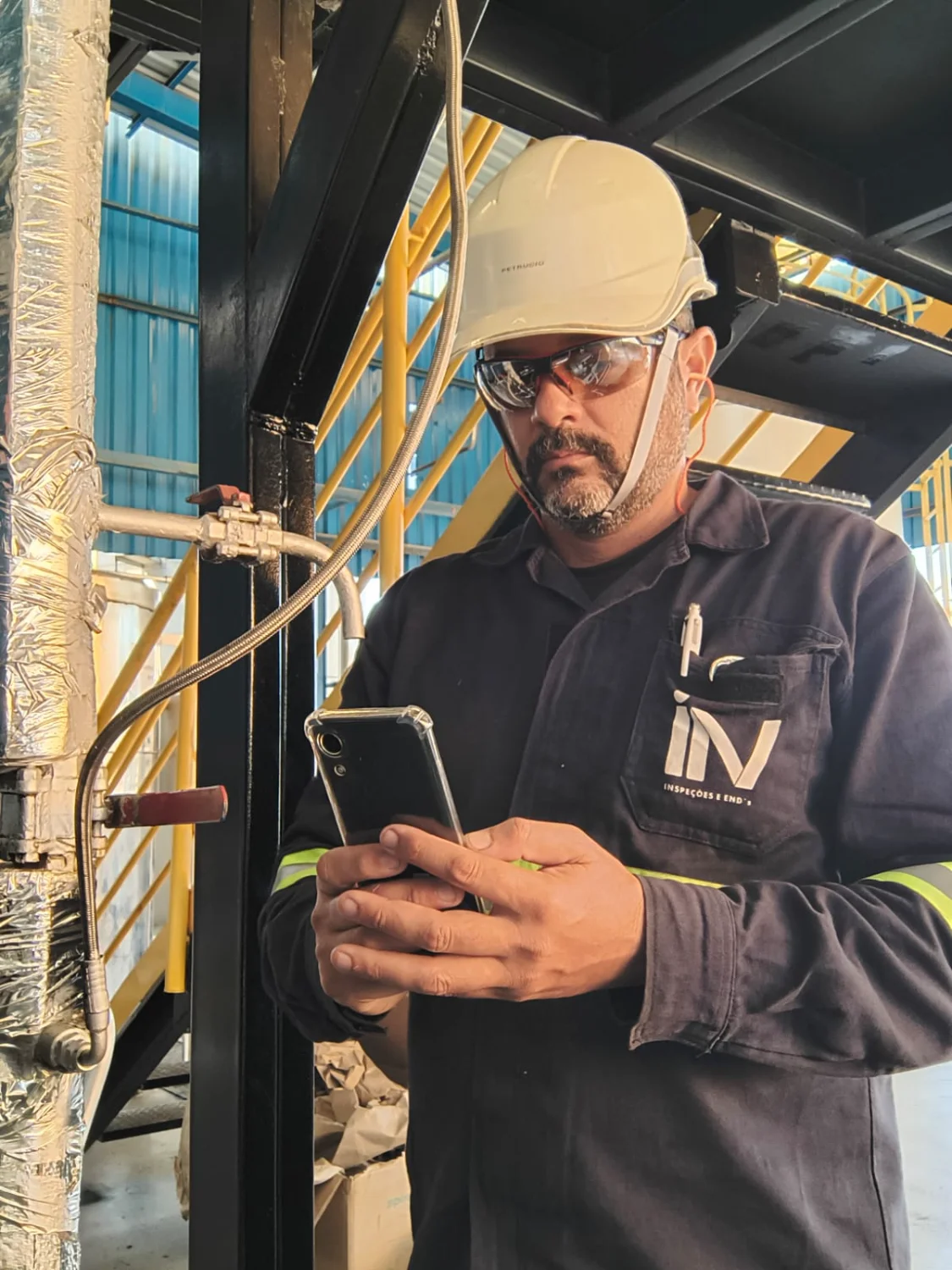 Engenheiro da Microsenior/INV com EPIs, usando smartphone para registro durante inspeção em tubulação industrial.