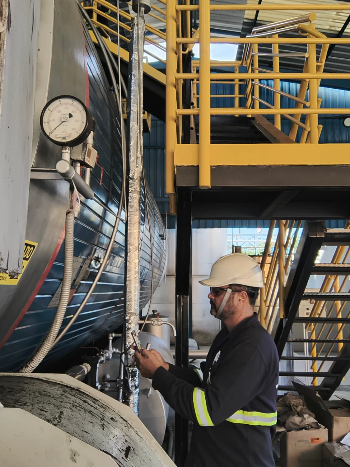 Técnico da INV com capacete e óculos, realizando calibração em manômetro de um vaso de pressão industrial.