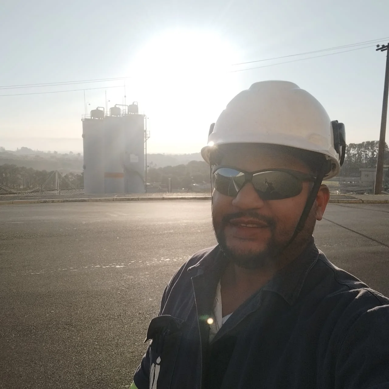 Engenheiro sorrindo, usando capacete e óculos em campo industrial com reservatório ao fundo.